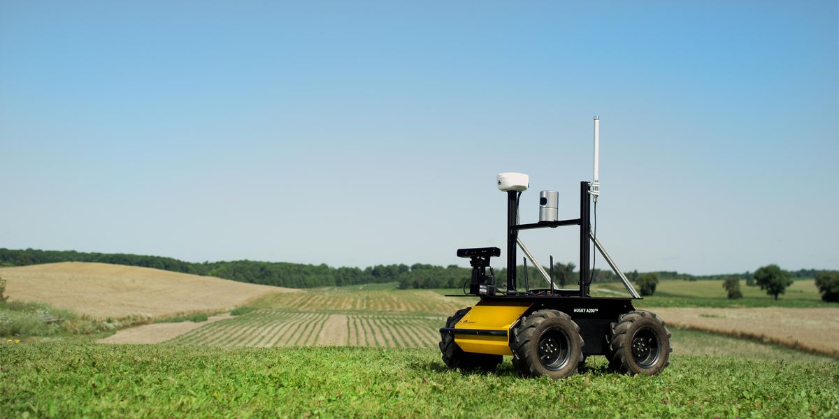 A photo of a small yellow and black wheeled robot with sensors on top on grass with rolling fields in the background.